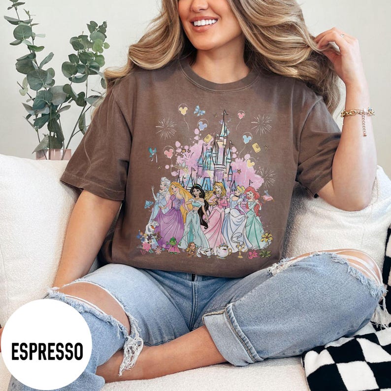 May include: Espresso-colored t-shirt featuring a colorful graphic of Disney princesses in front of a castle. The design includes fireworks, butterflies, and floral elements. The shirt is a relaxed fit.