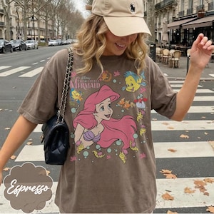 May include: Espresso-coloured t-shirt featuring a graphic of Ariel from Disney's The Little Mermaid. The design includes Ariel with pink hair, Flounder, and other sea creatures. The shirt has a relaxed fit.