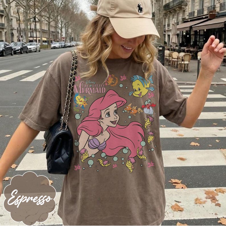 May include: Espresso-coloured t-shirt featuring a graphic of Ariel from Disney's The Little Mermaid. The design includes Ariel with pink hair, Flounder, and other sea creatures. The shirt has a relaxed fit.