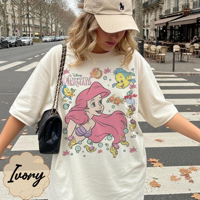 May include: Ivory t-shirt with a graphic of Ariel from Disney's The Little Mermaid. The design features Ariel with pink hair, a purple seashell bra, and sea creatures. The shirt has short sleeves and a crew neck.