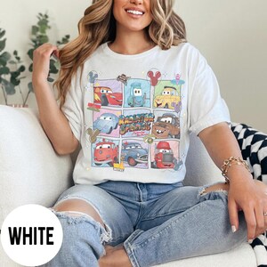 May include: White t-shirt featuring a colorful graphic of cartoon cars in a grid pattern. The design includes the text "Radiator Springs" and various car characters. The shirt is a casual, unisex style.