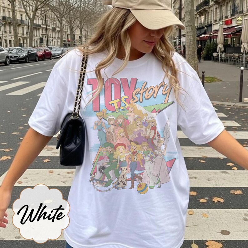 May include: A white short-sleeved t-shirt featuring a large graphic print with the text "TOY story" in pink and blue letters. The graphic depicts a group of female characters in various outfits, including a ball, against a colorful background.