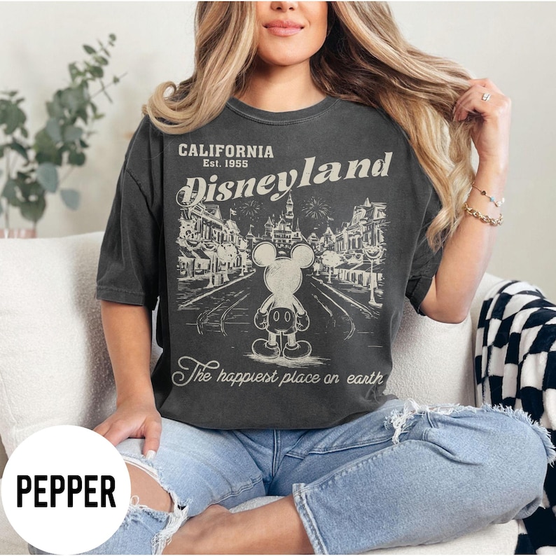 Może przedstawiać: Ciemnoszara koszulka z grafiką Disneyland. Grafika przedstawia Myszka Miki przed sceną uliczną z napisem "California Disneyland Est. 1955" i "The happiest place on earth."