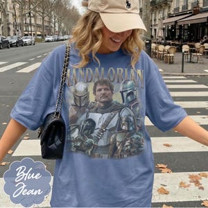 Pu&ograve; includere: Una t-shirt blu con una stampa grafica dei personaggi di The Mandalorian e il testo "Mandalorian" in lettere dorate. La maglietta ha maniche corte e scollo rotondo. &Egrave; indossata una borsa a tracolla nera trapuntata con una catena dorata.