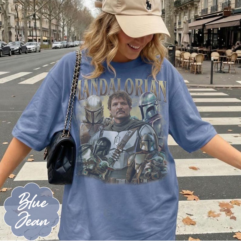 Pu&ograve; includere: Una t-shirt blu con una stampa grafica dei personaggi di The Mandalorian e il testo "Mandalorian" in lettere dorate. La maglietta ha maniche corte e scollo rotondo. &Egrave; indossata una borsa a tracolla nera trapuntata con una catena dorata.