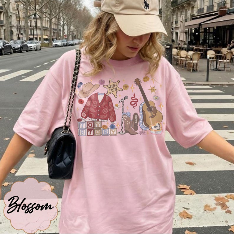 May include: A light pink t-shirt featuring a graphic print of a cowboy hat, a paisley cardigan, a sheriff badge labeled "Sheriff Taylor," a microphone, a snake, a guitar, cowboy boots, and blocks spelling "Toy Story" with floral accents.