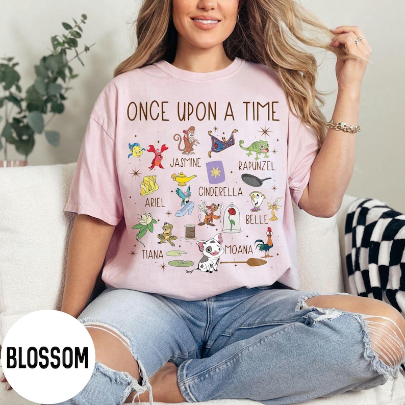 May include: Pale pink t-shirt featuring the words "ONCE UPON A TIME" and illustrations of cartoon characters such as Ariel, Jasmine, and Moana. The shirt is a light pink colour.