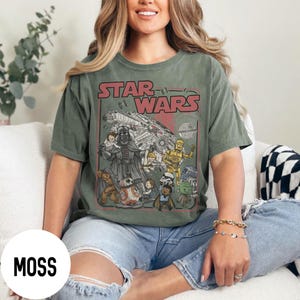 May include: Moss green Star Wars t-shirt featuring a vintage-style graphic with Darth Vader, the Millennium Falcon, and other characters. The text "STAR WARS" is in red. The shirt is a short-sleeved, crew neck style.