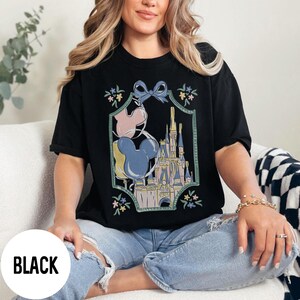 Może przedstawiać: Czarny t-shirt z kolorową grafiką zamku, balon&oacute;w Myszki Miki i kwiatowych akcent&oacute;w. Koszulka ma luźny kr&oacute;j i jest wykonana z miękkiego materiału. Słowo "BLACK" jest wydrukowane w lewym dolnym rogu obrazu.