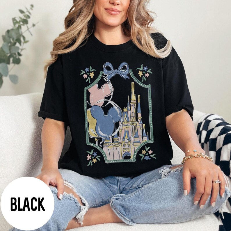 Może przedstawiać: Czarny t-shirt z kolorową grafiką zamku, balon&oacute;w Myszki Miki i kwiatowych akcent&oacute;w. Koszulka ma luźny kr&oacute;j i jest wykonana z miękkiego materiału. Słowo "BLACK" jest wydrukowane w lewym dolnym rogu obrazu.