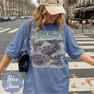 May include: A blue denim-colored t-shirt featuring a graphic print of multiple Grogu characters and the text GROGU at the top. The shirt has short sleeves and a crew neckline. A black quilted shoulder bag with a gold chain strap is worn over one shoulder.