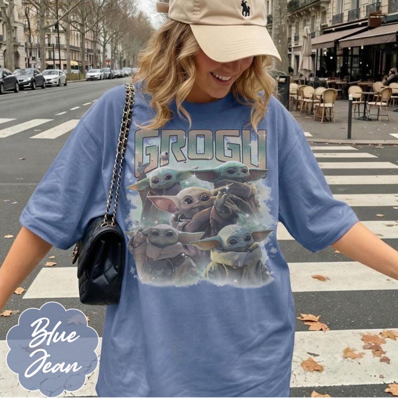 May include: A blue denim-colored t-shirt featuring a graphic print of multiple Grogu characters and the text GROGU at the top. The shirt has short sleeves and a crew neckline. A black quilted shoulder bag with a gold chain strap is worn over one shoulder.