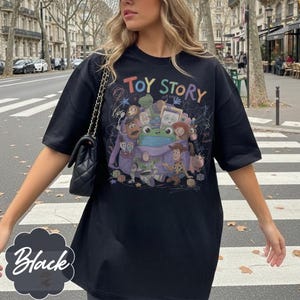May include: A black short-sleeve t-shirt featuring a colourful graphic print of Toy Story characters emerging from a purple backpack. The design includes the text "TOY STORY" at the top, "yee haw" written in script, and various scribbled doodle accents.
