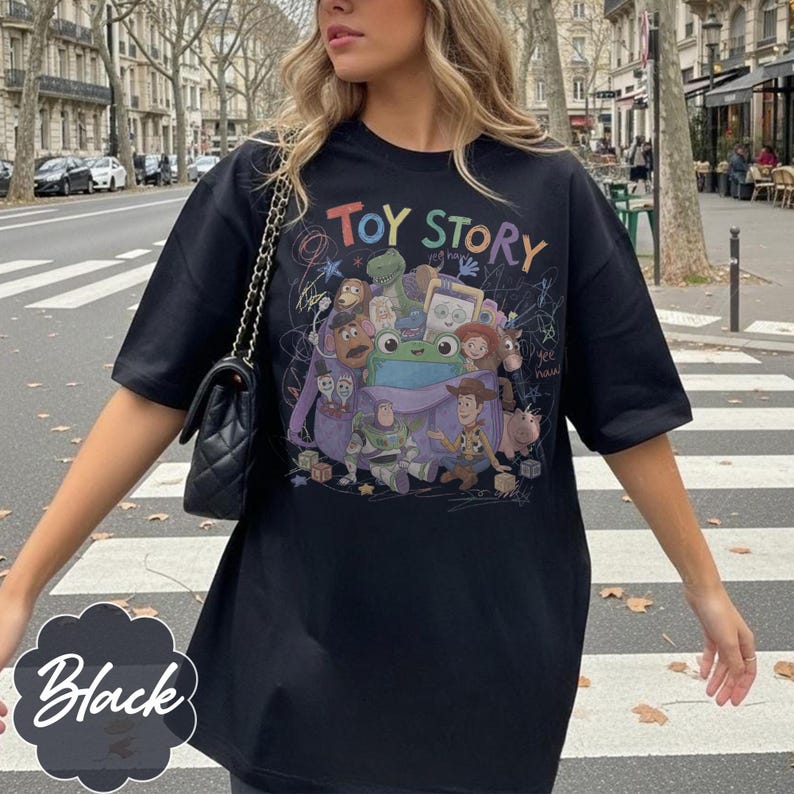 May include: A black short-sleeve t-shirt featuring a colourful graphic print of Toy Story characters emerging from a purple backpack. The design includes the text "TOY STORY" at the top, "yee haw" written in script, and various scribbled doodle accents.