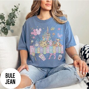 May include: A blue t-shirt with a colorful graphic featuring Disney characters, a castle, and the word "Disneyworld". The design includes pink balloons, popcorn, and a bow. The shirt is a comfortable, casual style.