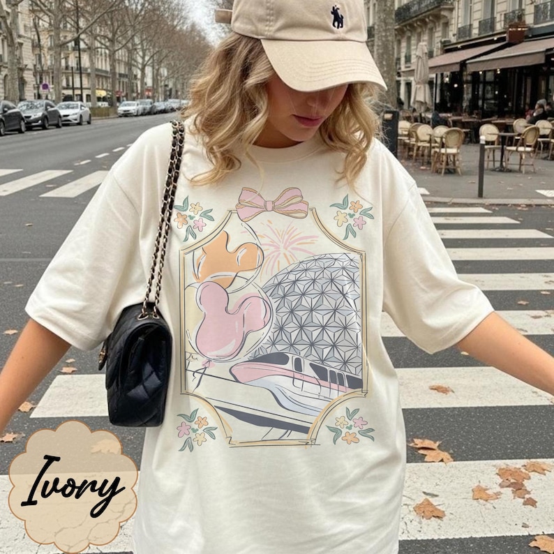 May include: Ivory-colored t-shirt featuring a colorful graphic of a theme park with Mickey Mouse. The design includes a pink bow, fireworks, and floral accents. The word "Ivory" is written in cursive at the bottom.