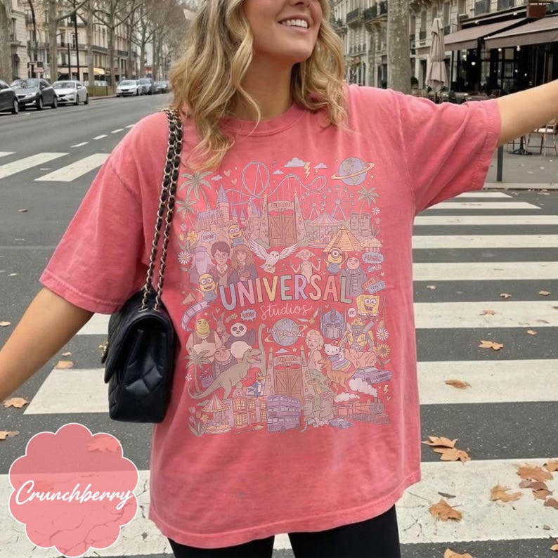 May include: A coral-coloured t-shirt with a vibrant graphic of Universal Studios characters and attractions. The text "UNIVERSAL Studios" is visible. The shirt has short sleeves and a relaxed fit, suitable for everyday wear.
