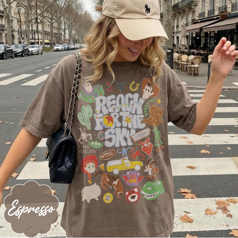 Pode incluir: Camiseta cor de caf&eacute; com um design gr&aacute;fico com personagens e a frase "Reach for the Sky". O design inclui um cacto, um cowboy, um guarda florestal espacial e outros elementos de desenho animado. A camisa &eacute; combinada com um bon&eacute; bege e uma bolsa preta.