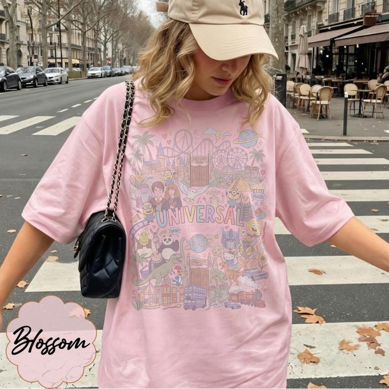 May include: A pale pink t-shirt featuring a detailed illustration of a theme park, with the word "UNIVERSAL" printed across the design. The shirt has short sleeves and a loose fit. A black shoulder bag is visible.