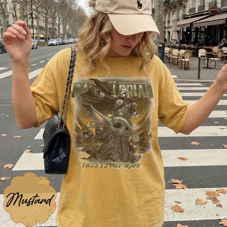 Puede incluir: Una camiseta color amarillo mostaza con un estampado gr&aacute;fico de The Mandalorian y Grogu. El texto dice "The Mandalorian" y "This is the way". La camiseta tiene mangas cortas y cuello redondo, acompa&ntilde;ada de un bolso de hombro negro acolchado.