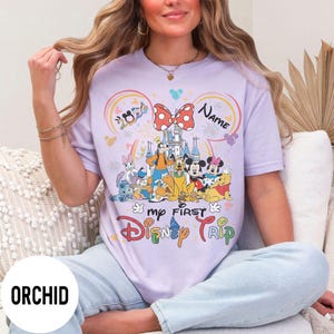 May include: Orchid purple t-shirt featuring a colorful Disney graphic. The design includes Disney characters, a castle, and the text "my FIRST Disney TRIP". The shirt has a crew neck and short sleeves.