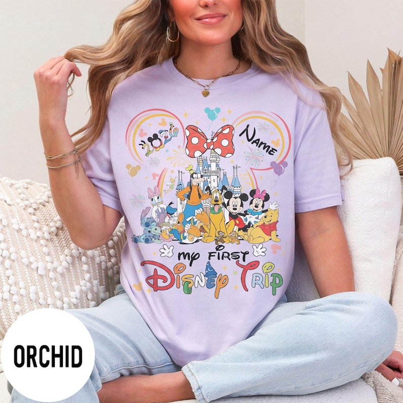 May include: Orchid purple t-shirt featuring a colorful Disney graphic. The design includes Disney characters, a castle, and the text "my FIRST Disney TRIP". The shirt has a crew neck and short sleeves.