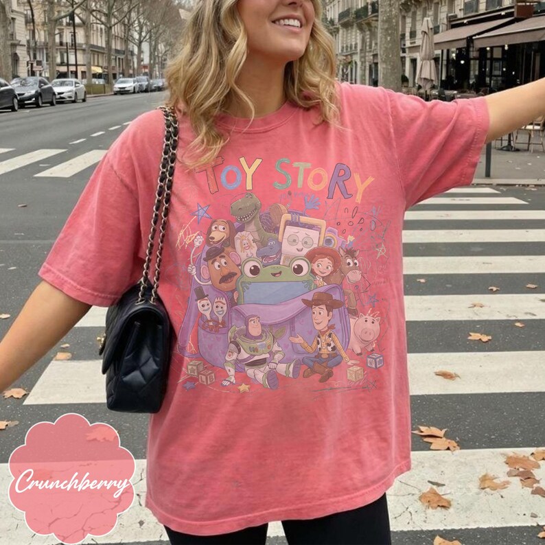 May include: A coral-coloured short-sleeve t-shirt featuring a graphic print of Toy Story characters including Woody, Buzz Lightyear, and a green frog. The text Toy Story is printed in colourful letters above the characters. A black quilted shoulder bag.