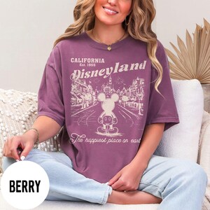 Może przedstawiać: Koszulka w kolorze jagodowym z grafiką Disneyland. Projekt zawiera napis "CALIFORNIA Est. 1955 Disneyland" z ilustracją Myszki Miki i frazą "The happiest place on earth."
