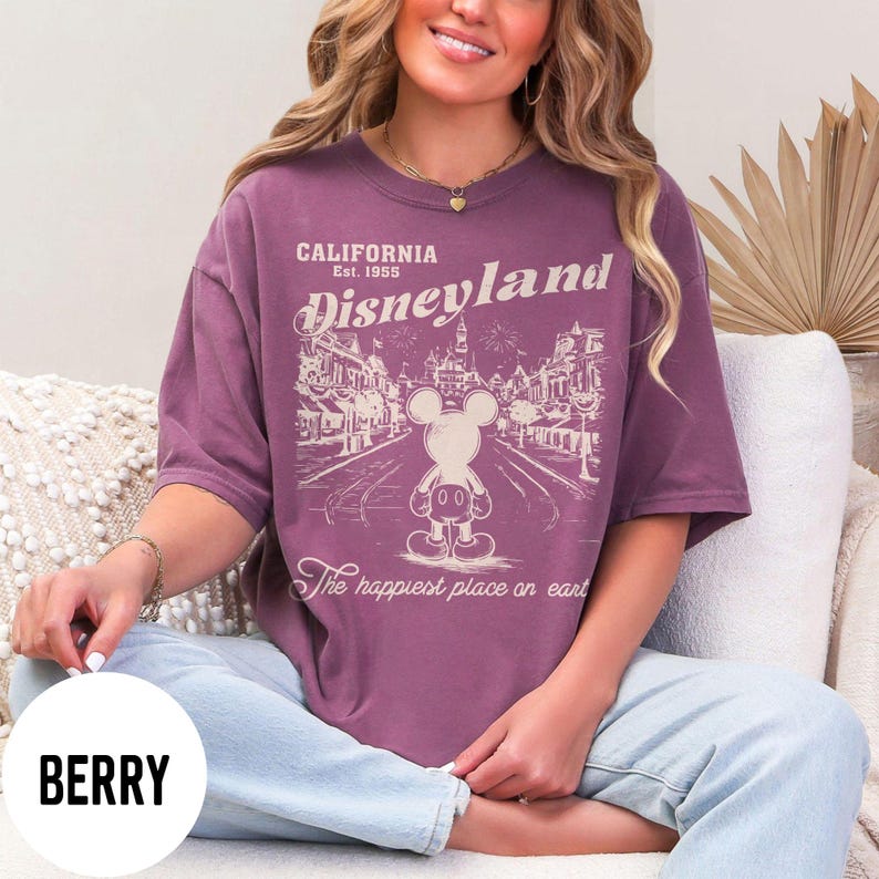 Może przedstawiać: Koszulka w kolorze jagodowym z grafiką Disneyland. Projekt zawiera napis "CALIFORNIA Est. 1955 Disneyland" z ilustracją Myszki Miki i frazą "The happiest place on earth."