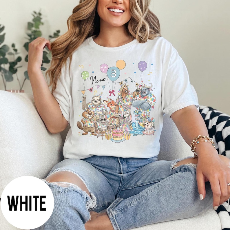 以下が含まれることがあります： 誕生日を祝う擬人化された動物たちのカラフルなイラストが描かれた白いTシャツ。デザインには風船、ケーキ、数字の3が含まれています。「Name」という文字も表示されています。