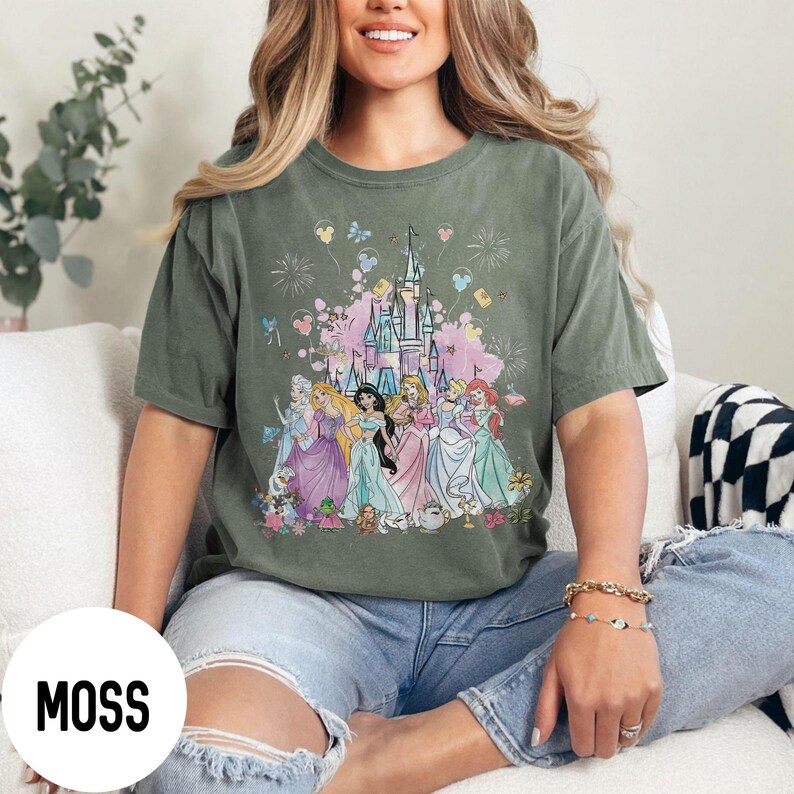 May include: Moss green t-shirt featuring a colorful graphic of a castle and several princesses. The princesses are wearing gowns in shades of pink, blue, and purple. The word "MOSS" is printed in white on a white circle at the bottom left.