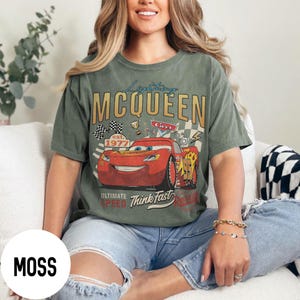 Op de afbeelding: Mosgroen t-shirt met een afbeelding van Bliksem McQueen uit de film Cars. Het ontwerp bevat de tekst "Lightning McQueen" en "Ultimate Speed Think Fast" met racevlaggen en trofee&euml;n. Het shirt heeft een relaxte pasvorm.