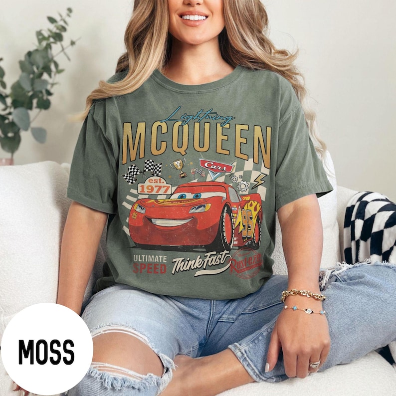 Op de afbeelding: Mosgroen t-shirt met een afbeelding van Bliksem McQueen uit de film Cars. Het ontwerp bevat de tekst "Lightning McQueen" en "Ultimate Speed Think Fast" met racevlaggen en trofee&euml;n. Het shirt heeft een relaxte pasvorm.