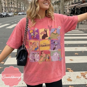 May include: A coral-colored short-sleeve t-shirt featuring a grid of colorful cartoon characters from Disney's The Emperor's New Groove. The graphic includes Kuzco, Pacha, Yzma, and Kronk, with the movie title text centered in the middle square.