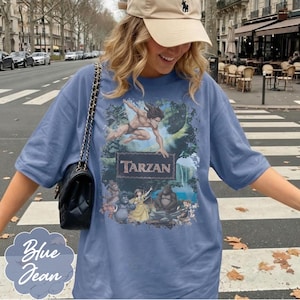 Op de afbeelding: Een blauw spijkerstof T-shirt met een Tarzan-afbeelding. De afbeelding toont Tarzan, junglebladeren en andere personages. Het shirt heeft korte mouwen en een relaxte pasvorm. De woorden "Blue Jean" staan in lichtblauw cursief.