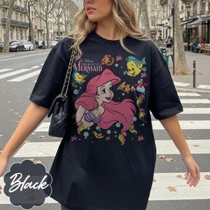 May include: Black t-shirt with a colourful graphic of Ariel from Disney's The Little Mermaid. The design includes Ariel, Flounder, and other sea creatures. The text "Disney The Little Mermaid" is also visible.