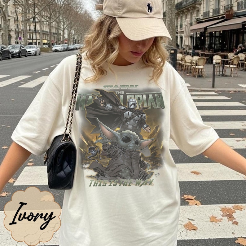 Puede incluir: Una camiseta de manga corta color marfil con un estampado gr&aacute;fico de The Mandalorian y Grogu de Star Wars. El dise&ntilde;o incluye el texto Star Wars, The Mandalorian y This Is The Way. Se lleva un bolso de hombro negro con correa de cadena.