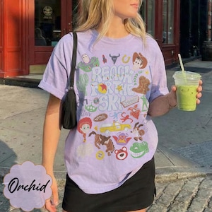 Pode incluir: Uma camiseta lil&aacute;s clara com um design gr&aacute;fico com personagens e frases do filme "Toy Story". A camiseta inclui a frase "Reach for the Sky" e v&aacute;rias imagens de desenhos animados. A camiseta &eacute; combinada com uma saia preta.
