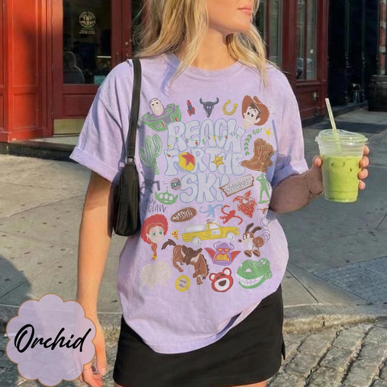 Pode incluir: Uma camiseta lil&aacute;s clara com um design gr&aacute;fico com personagens e frases do filme "Toy Story". A camiseta inclui a frase "Reach for the Sky" e v&aacute;rias imagens de desenhos animados. A camiseta &eacute; combinada com uma saia preta.