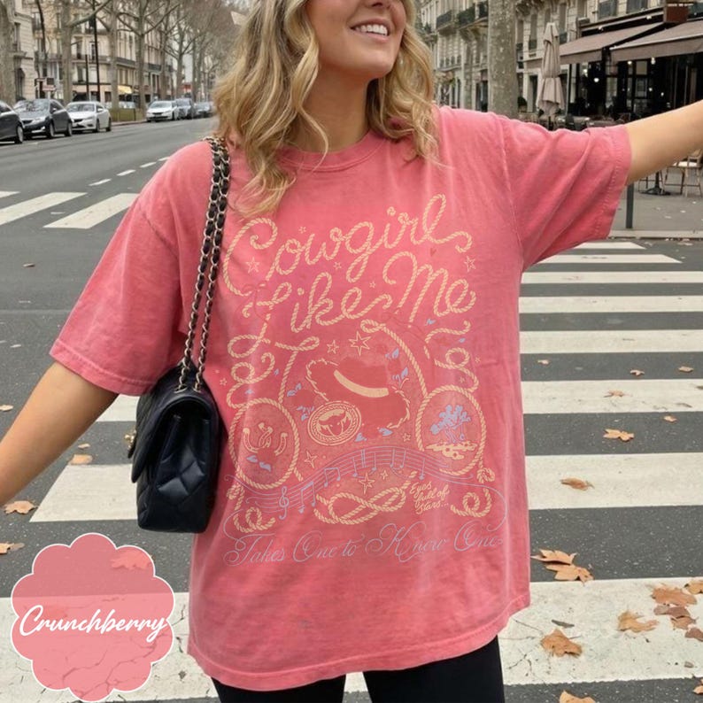 May include: A coral-colored short-sleeve t-shirt featuring a graphic print in light yellow and blue. The design includes the text "Cowgirl Like Me" and "Takes One to Know One" with illustrations of a cowboy hat, rope, and musical notes.