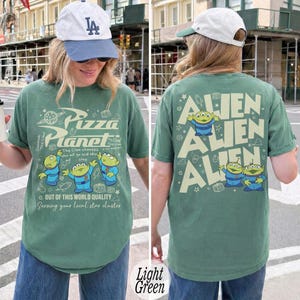 K&ouml;nnte beinhalten: Hellgr&uuml;nes T-Shirt mit einem Pizza Planet-Motiv, das den Text "Pizza Planet" und Cartoon-Aliens zeigt. Auf der R&uuml;ckseite des Shirts steht das Wort "ALIEN" dreimal wiederholt mit Alien-Charakteren.