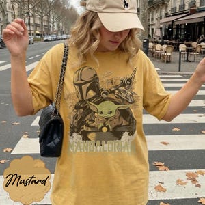 Może przedstawiać: Musztardowa koszulka z nadrukiem graficznym przedstawiającym The Mandalorian i Grogu z serii Star Wars. Projekt zawiera napis "Mandalorian". Koszulka ma luźny kr&oacute;j.
