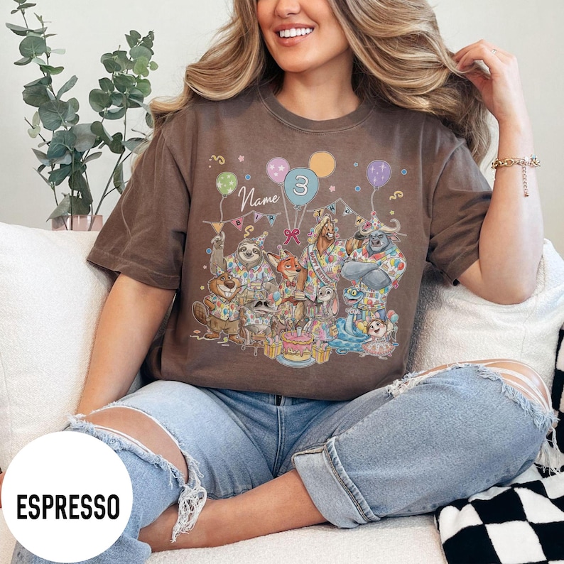 以下が含まれることがあります： エスプレッソ色のTシャツ。風船とケーキのある、カラフルな漫画の動物の誕生日パーティーのシーンが描かれています。風船には数字の3が描かれています。「Name」の文字が漫画のキャラクターの上にあります。ゆったりとしたフィット感のシャツです。