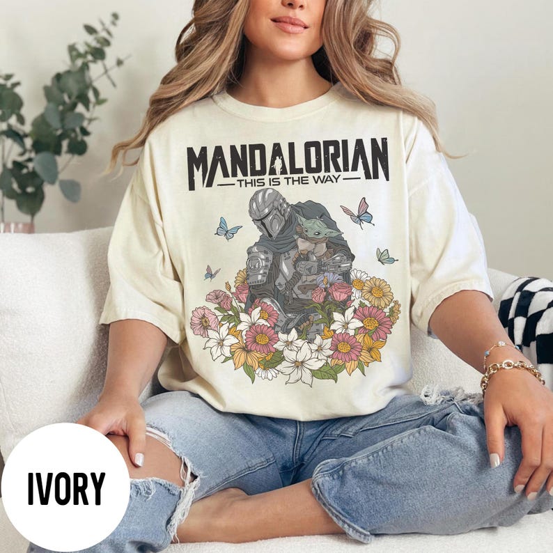 Puede incluir: Camiseta color marfil con el texto "MANDALORIAN" sobre una ilustraci&oacute;n del Mandaloriano y Grogu, rodeados de flores y mariposas. La frase "THIS IS THE WAY" est&aacute; debajo.