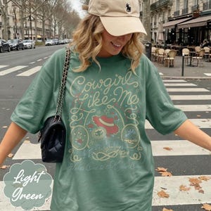 May include: A sage green short-sleeve t-shirt featuring a graphic print with the text Cowgirls Like Me and Takes One to Know One. The design includes a red hat, rope motifs, musical notes, and small floral elements in a light cream color.