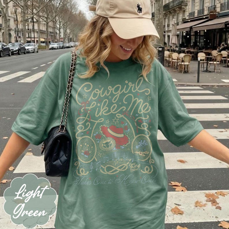 May include: A sage green short-sleeve t-shirt featuring a graphic print with the text Cowgirls Like Me and Takes One to Know One. The design includes a red hat, rope motifs, musical notes, and small floral elements in a light cream color.