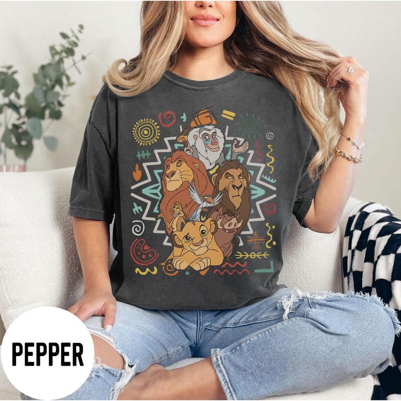 Puede incluir: Camiseta gris oscuro con un gr&aacute;fico colorido de personajes de El Rey Le&oacute;n. El dise&ntilde;o incluye leones, un suricato, un jabal&iacute; y otros personajes, con patrones de estilo tribal. La palabra "PEPPER" est&aacute; impresa en la parte inferior izquierda.