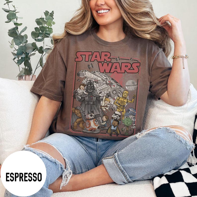 May include: Espresso-colored t-shirt featuring a Star Wars graphic. The design includes Darth Vader, C-3PO, R2-D2, and other characters, with the words "STAR WARS" in red. The shirt is worn with blue jeans.