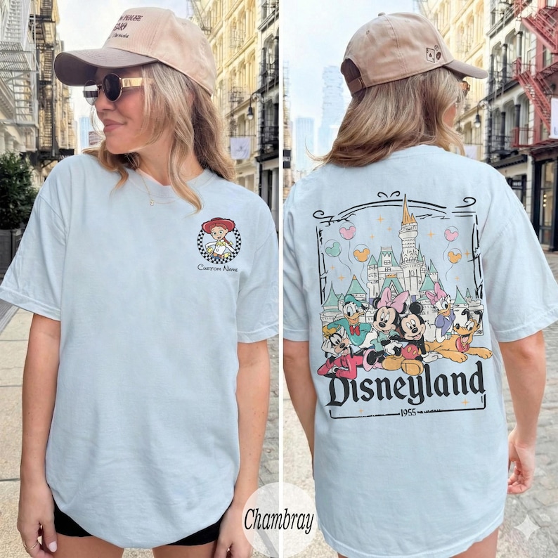 Pu&ograve; includere: T-shirt azzurra con tema Disney. La parte anteriore presenta un'immagine a cartoni animati di un personaggio e la scritta "Custom Name". La parte posteriore presenta una grafica di Disneyland con personaggi e la scritta "Disneyland 1955".