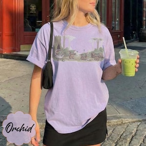 Puede incluir: Una camiseta de color lila claro con una ilustraci&oacute;n de dibujos animados de Grogu de The Mandalorian. El gr&aacute;fico muestra a Grogu en un entorno forestal. La palabra "Orchid" est&aacute; impresa en una etiqueta decorativa. La persona sostiene una bebida verde.